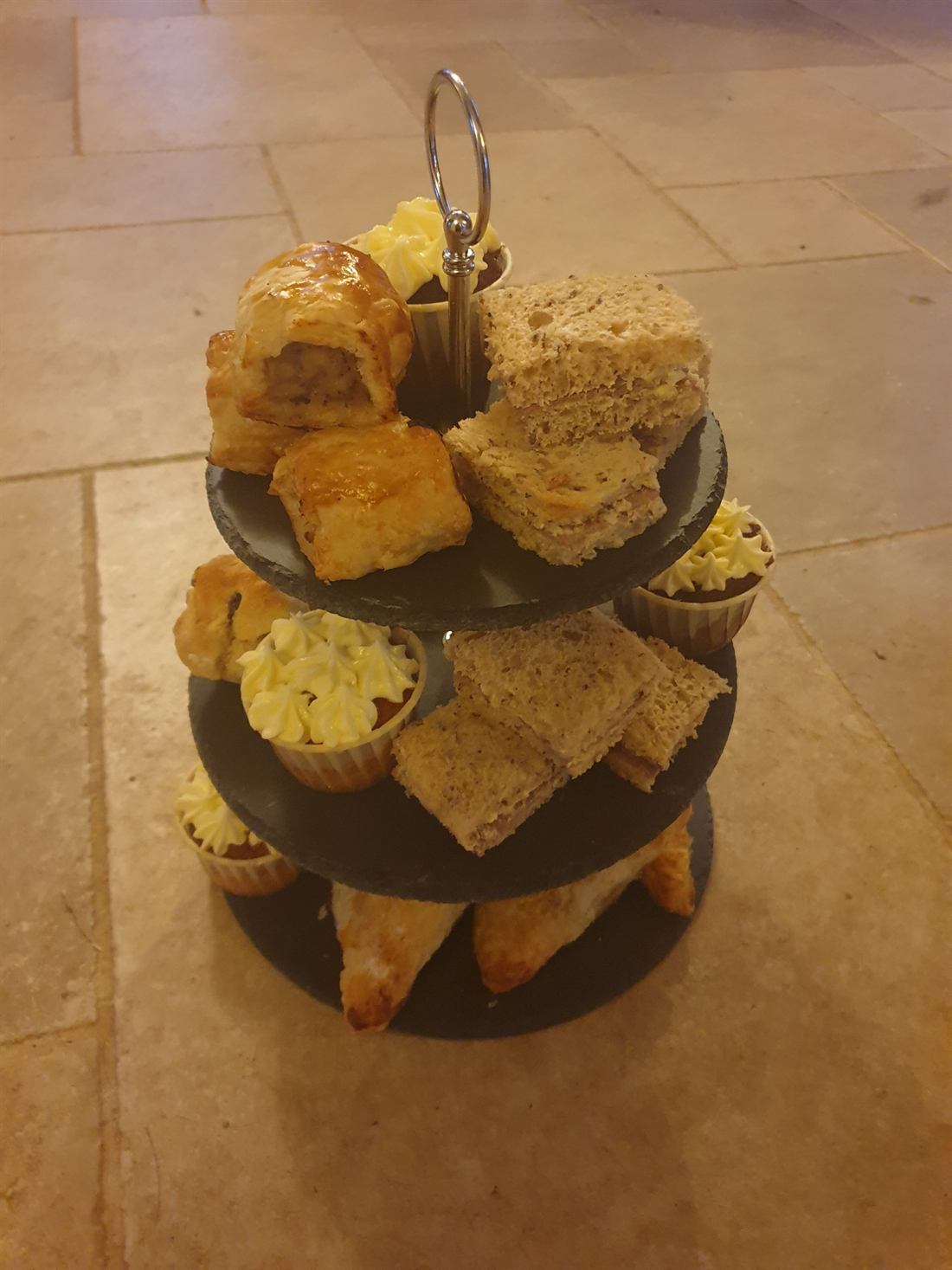 A three-tier slate stand of savoury pastries, sandwiches and cup cakes