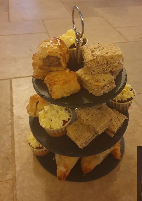 A three-tier slate stand of sausage rolls, sandwiches, scones and cup cakes